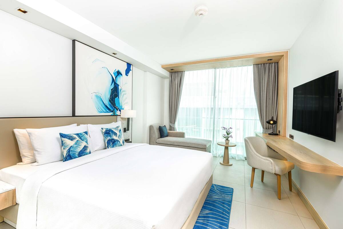 A hotel room with a large bed, a TV mounted on the wall, a seating area with a chair, and a balcony with curtains.