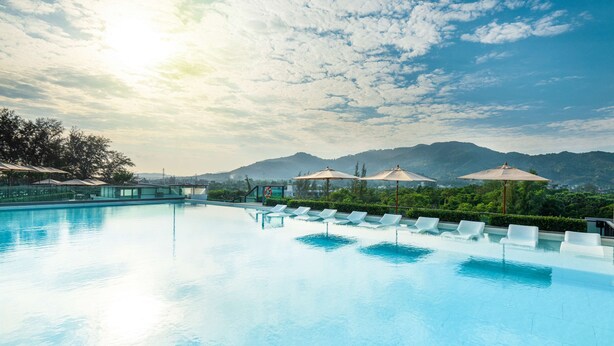 A swimming pool with lounge chairs and umbrellas, surrounded by greenery and mountains. - Hilton Garden Inn Phuket, Thailand (Choeng Thale)