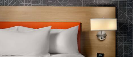 Una cama bien hecha con sábanas blancas y dos almohadas, cabecera de madera con un detalle naranja, una lámpara de noche y un reloj digital sobre una mesita de noche de madera.