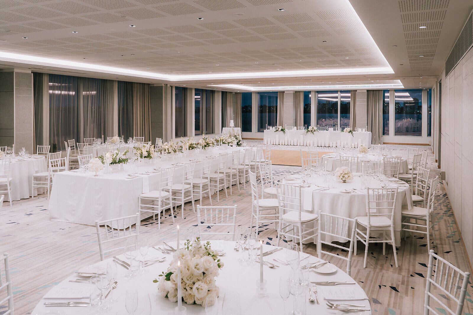 Une vaste salle de banquet, avec de nombreuses tables rondes dressées pour un événement formel, agrémentée de nappes blanches et de compositions florales.