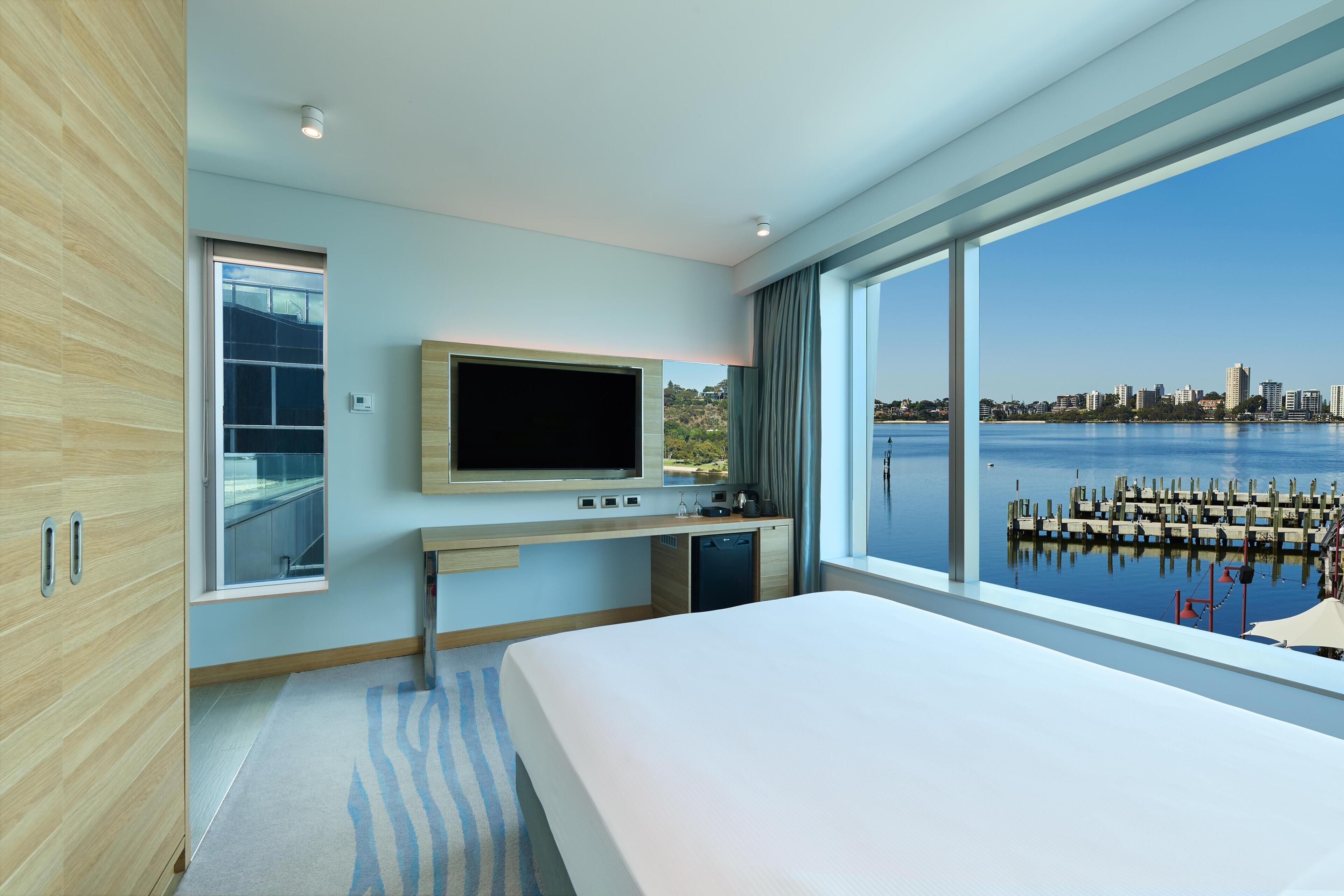 A modern hotel room with a large bed, a TV, a desk, and a view of a city skyline and water.