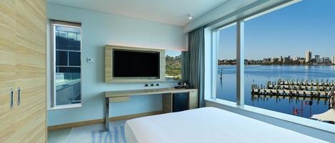 A modern hotel room with a large bed, a TV, a desk, and a view of a city skyline and water.