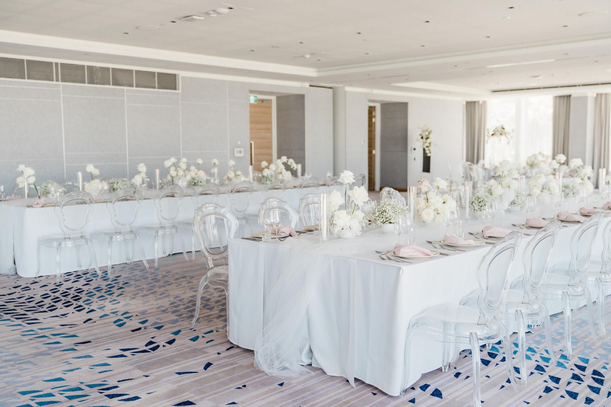 Une salle de banquet avec de longues tables dressées pour un événement formel, des chaises transparentes et des centres de table floraux.