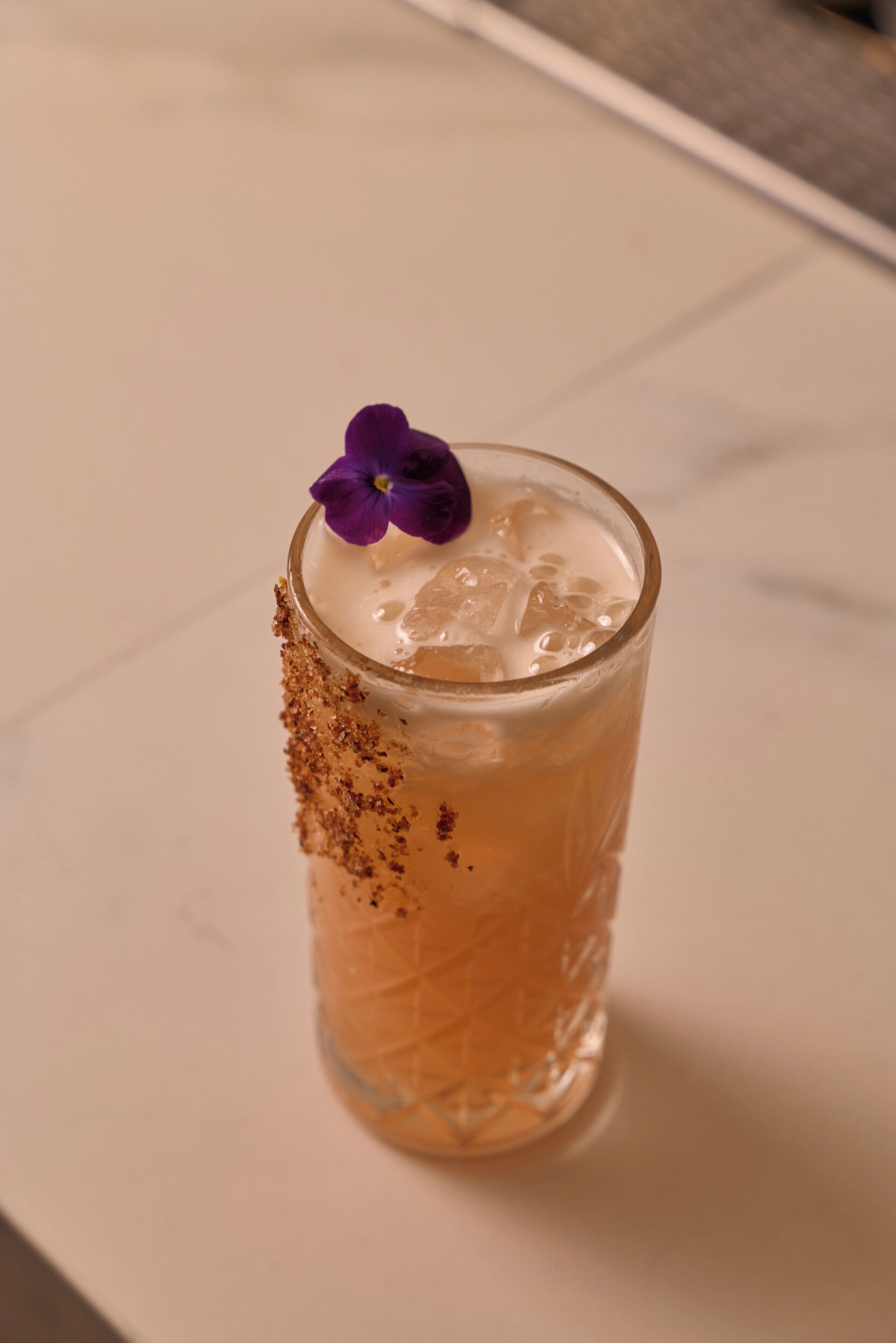A clear glass with a purple flower garnish and a sprinkle of brown powder on a white surface.