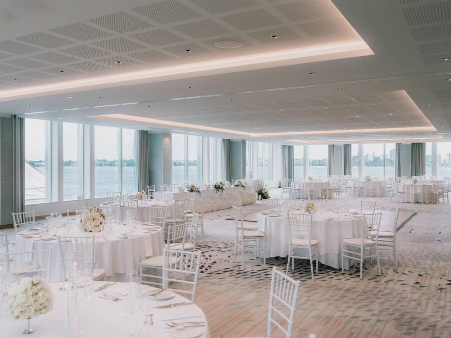 Un amplio salón de banquetes con mesas redondas dispuestas para comer, grandes ventanales con vistas al agua y un diseño de techo moderno.