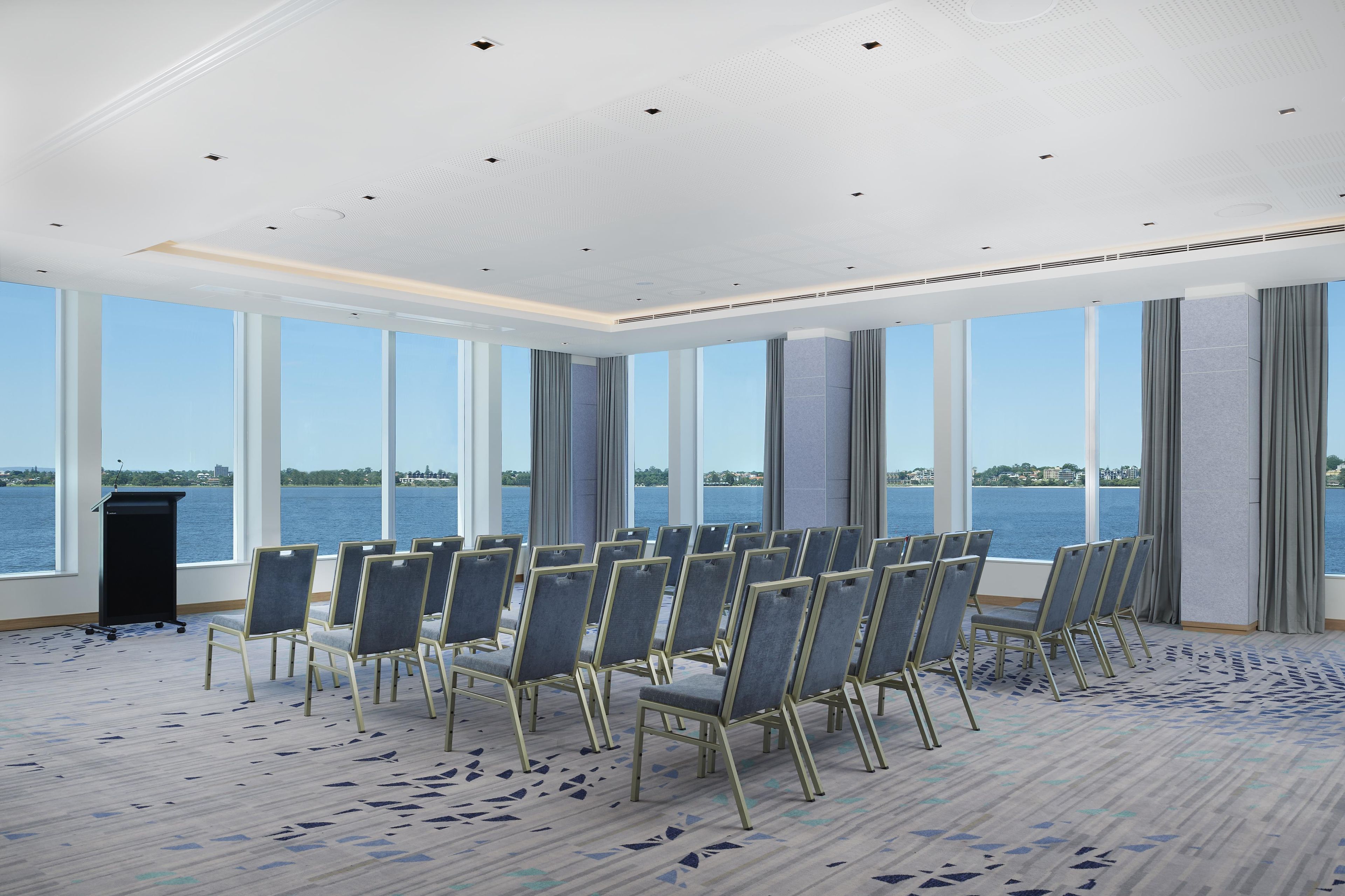 A conference room with a view of a body of water, rows of chairs, a podium, and a patterned carpet.