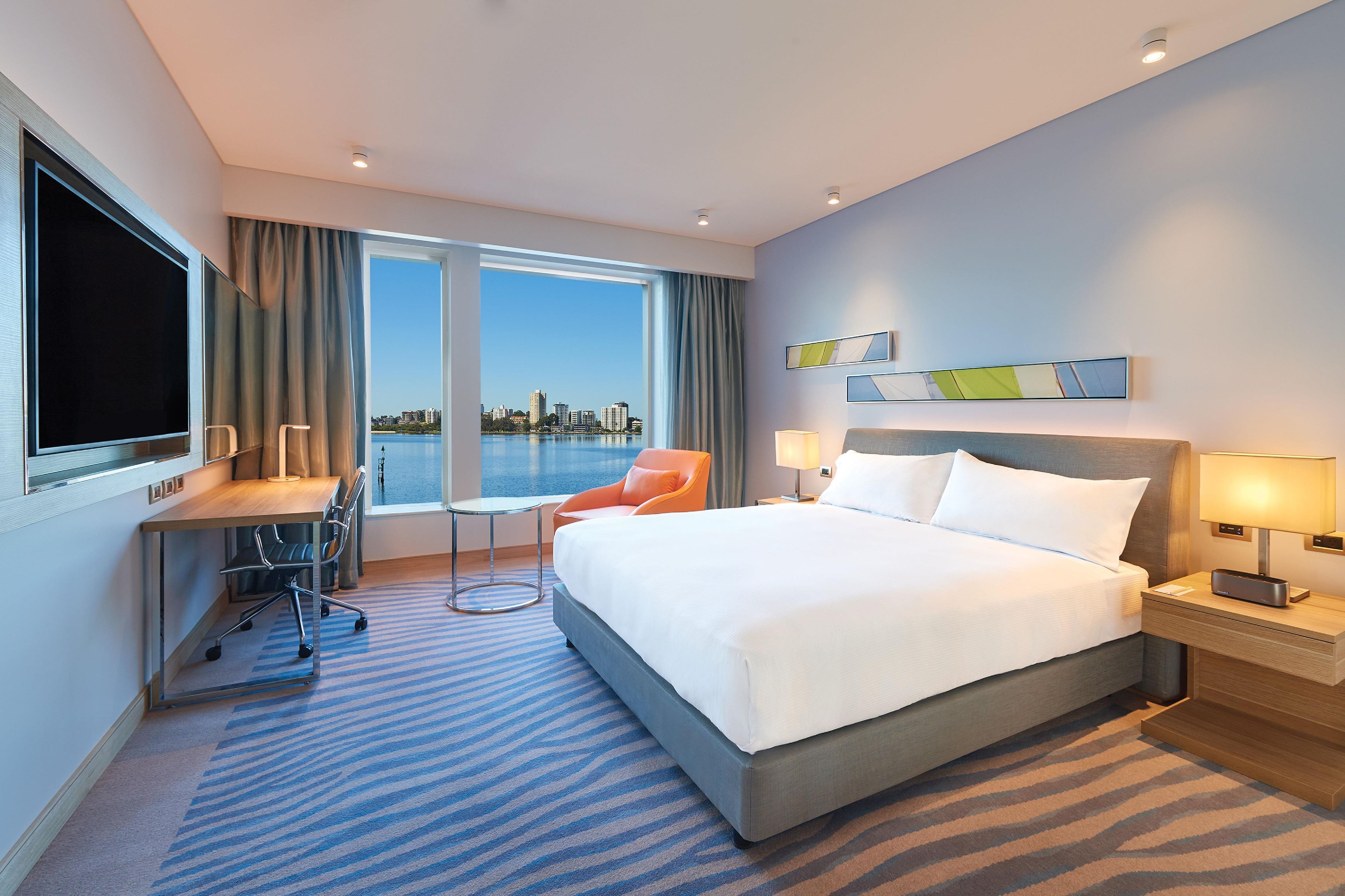 Habitación de hotel con cama, escritorio, silla, televisión y vistas a un paisaje urbano a través de amplias ventanas.