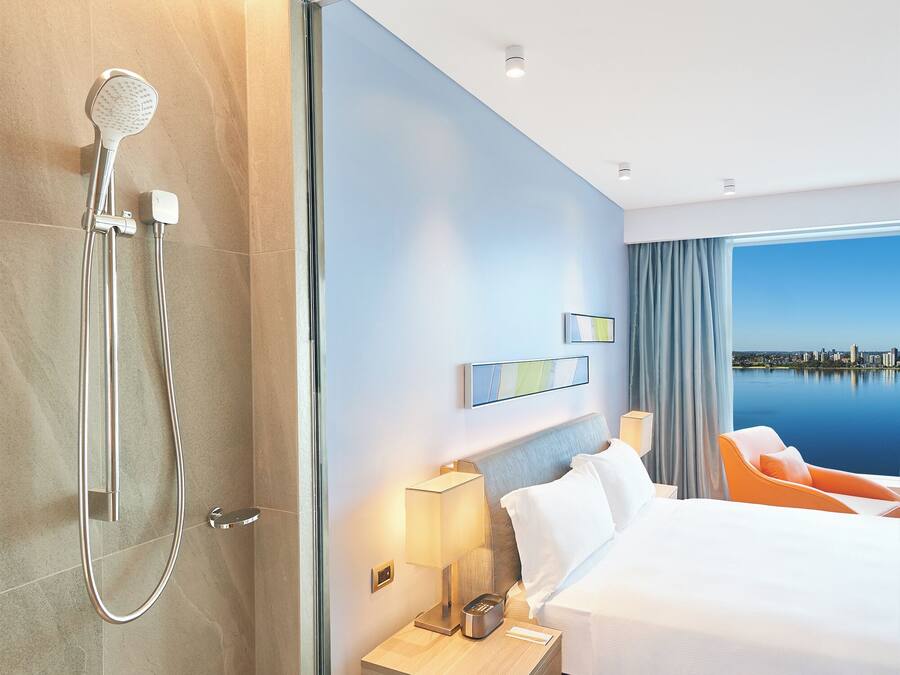 Una habitación de hotel moderna con una cama grande, una mesita de noche con lámpara, zona de ducha y vistas a un paisaje urbano a través de un ventanal amplio.