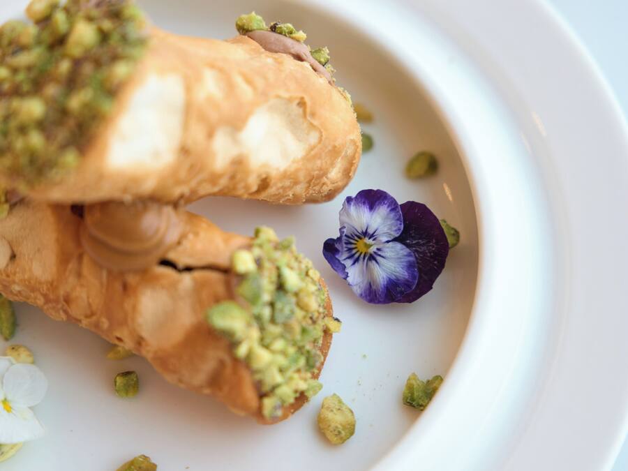 Un plato blanco con dos pastas cubiertas de pistachos verdes y una pequeña flor morada.