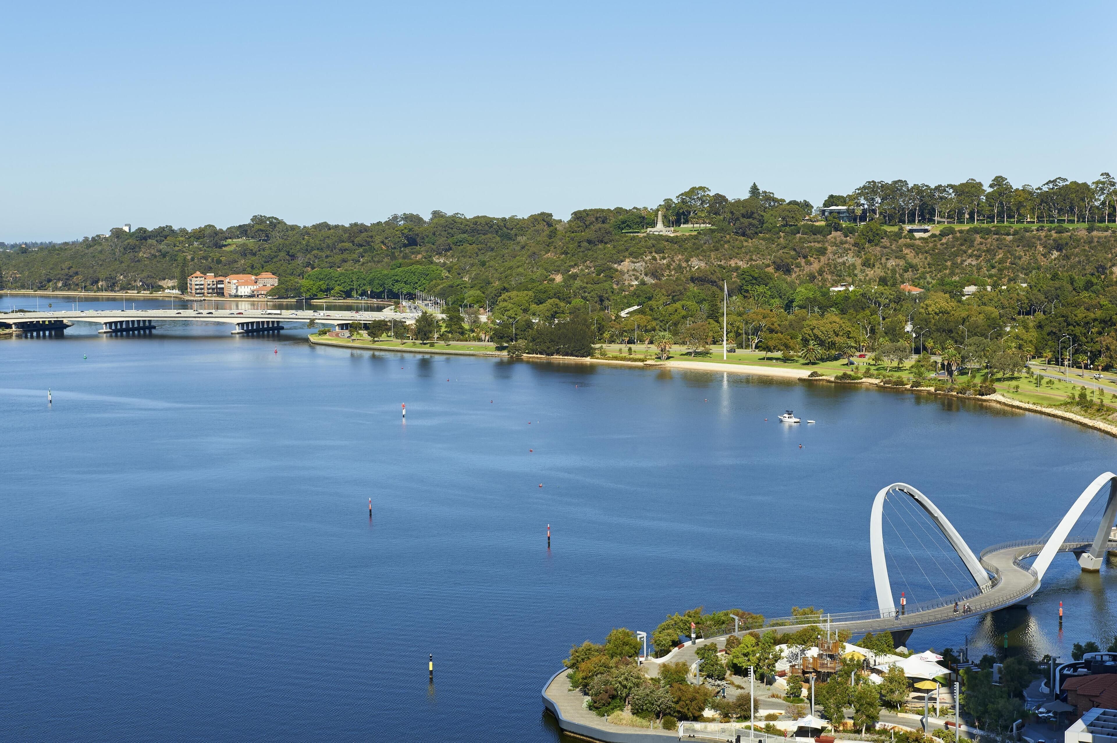 A large body of water with a modern bridge, a park with trees, and buildings in the background.