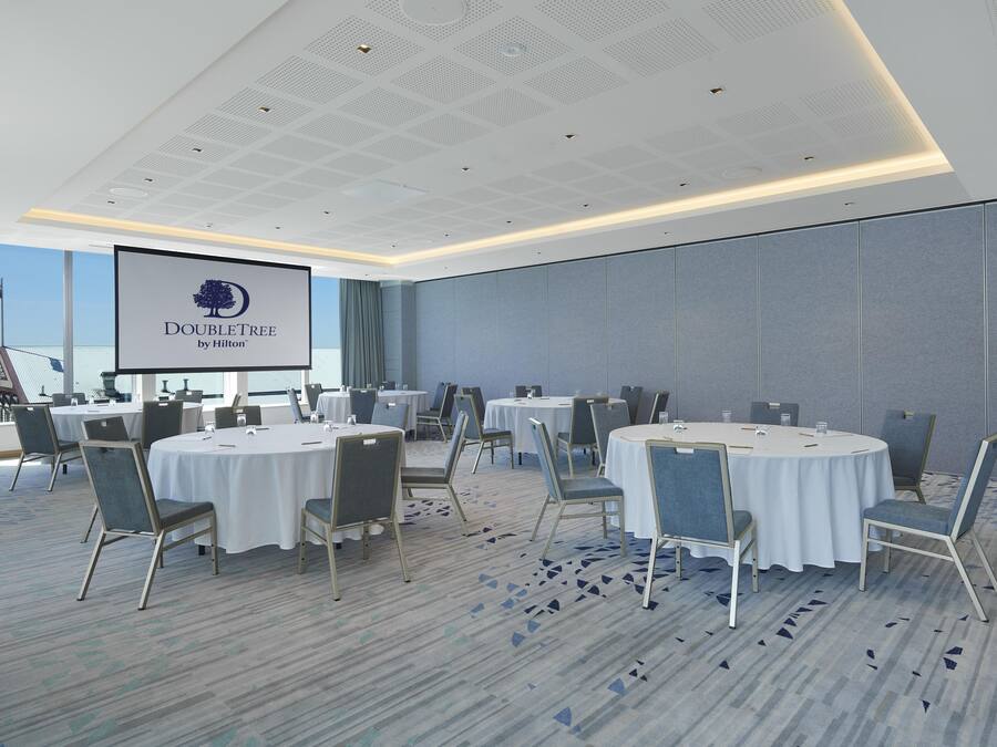 Un salón de conferencias con mesas redondas, sillas y una pantalla grande mostrando el logotipo de DoubleTree by Hilton.