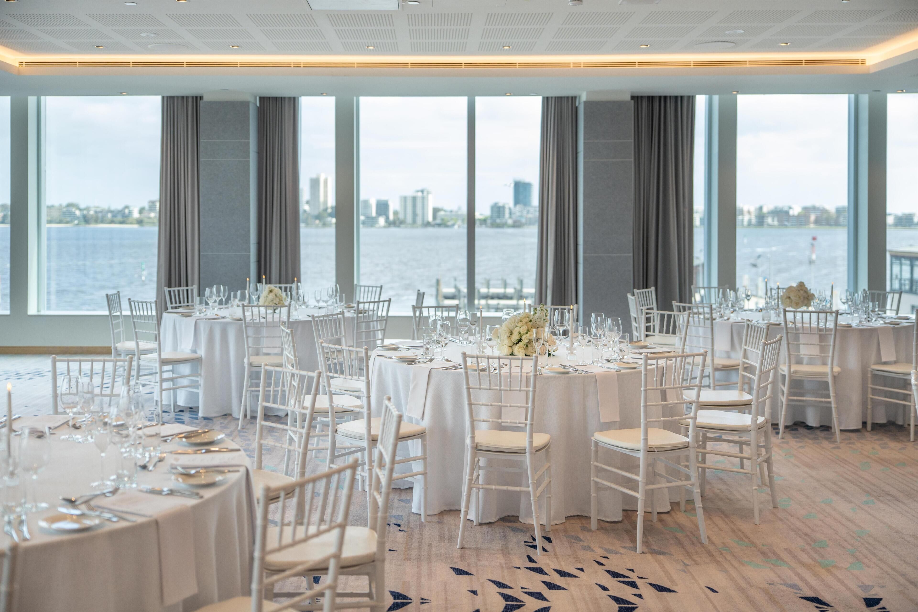 A banquet hall with round tables, white chairs, and a view of a body of water with a cityscape in the background.