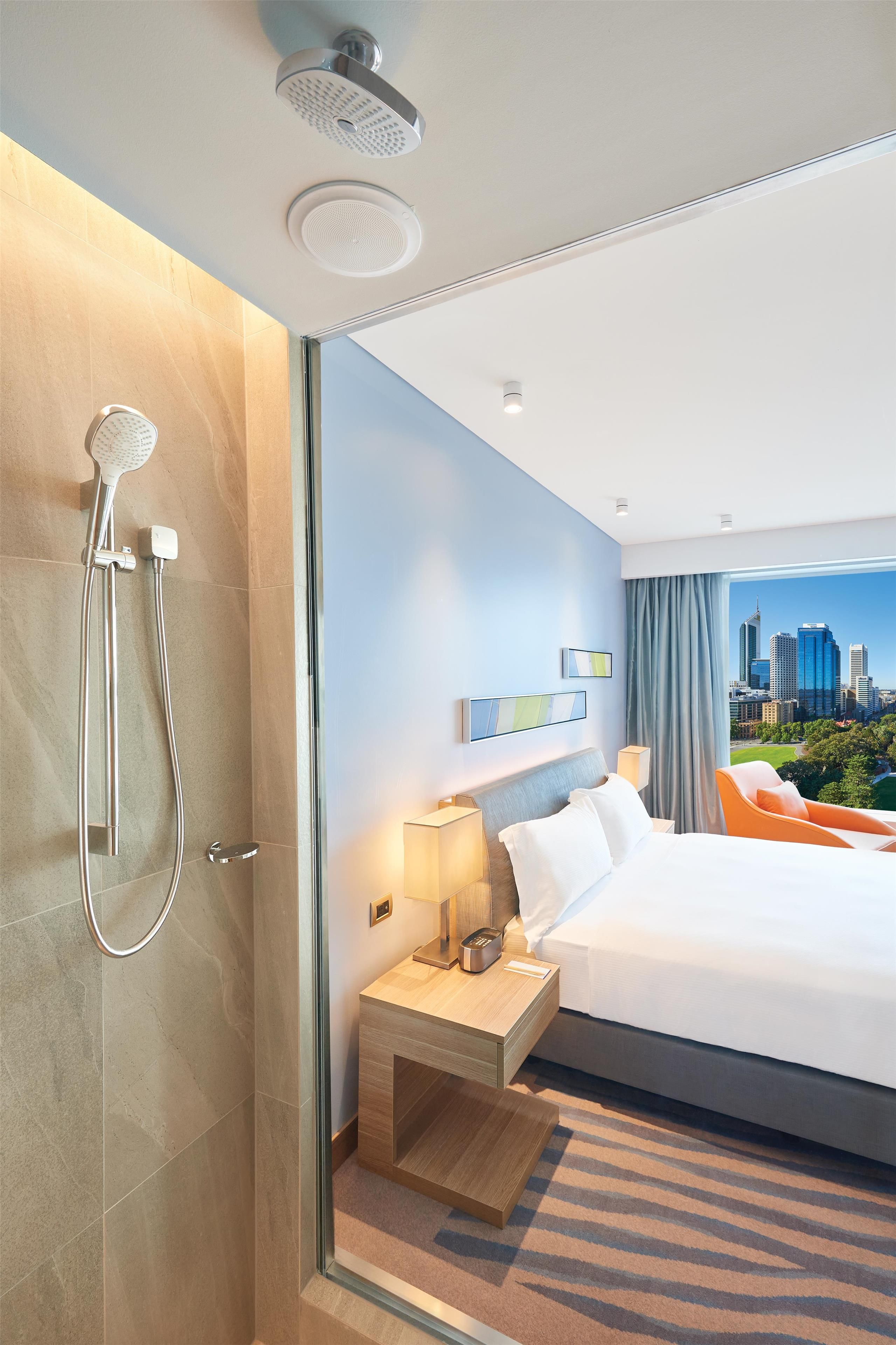 Quarto de hotel moderno com uma cama grande, mesa de cabeceira com abajur, chuveiro e vista para a cidade.