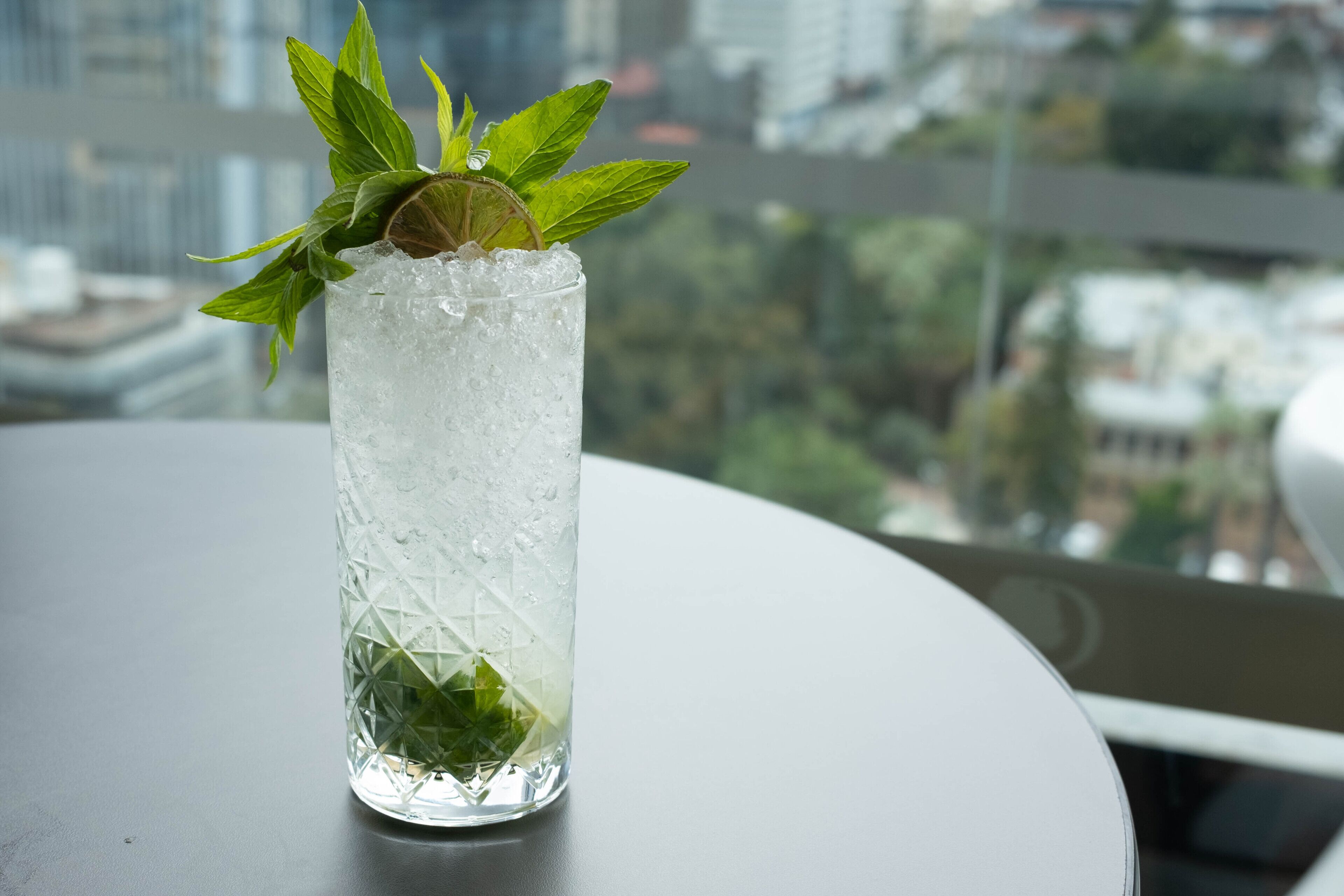 A tall glass with ice and mint leaves on a table with a city view in the background.