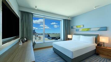 A hotel room with a bed, a desk, a TV, a window with a view, and a nightstand with a lamp.