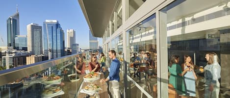 A dining area with a city skyline view, tables with food and drinks, and a glass railing.