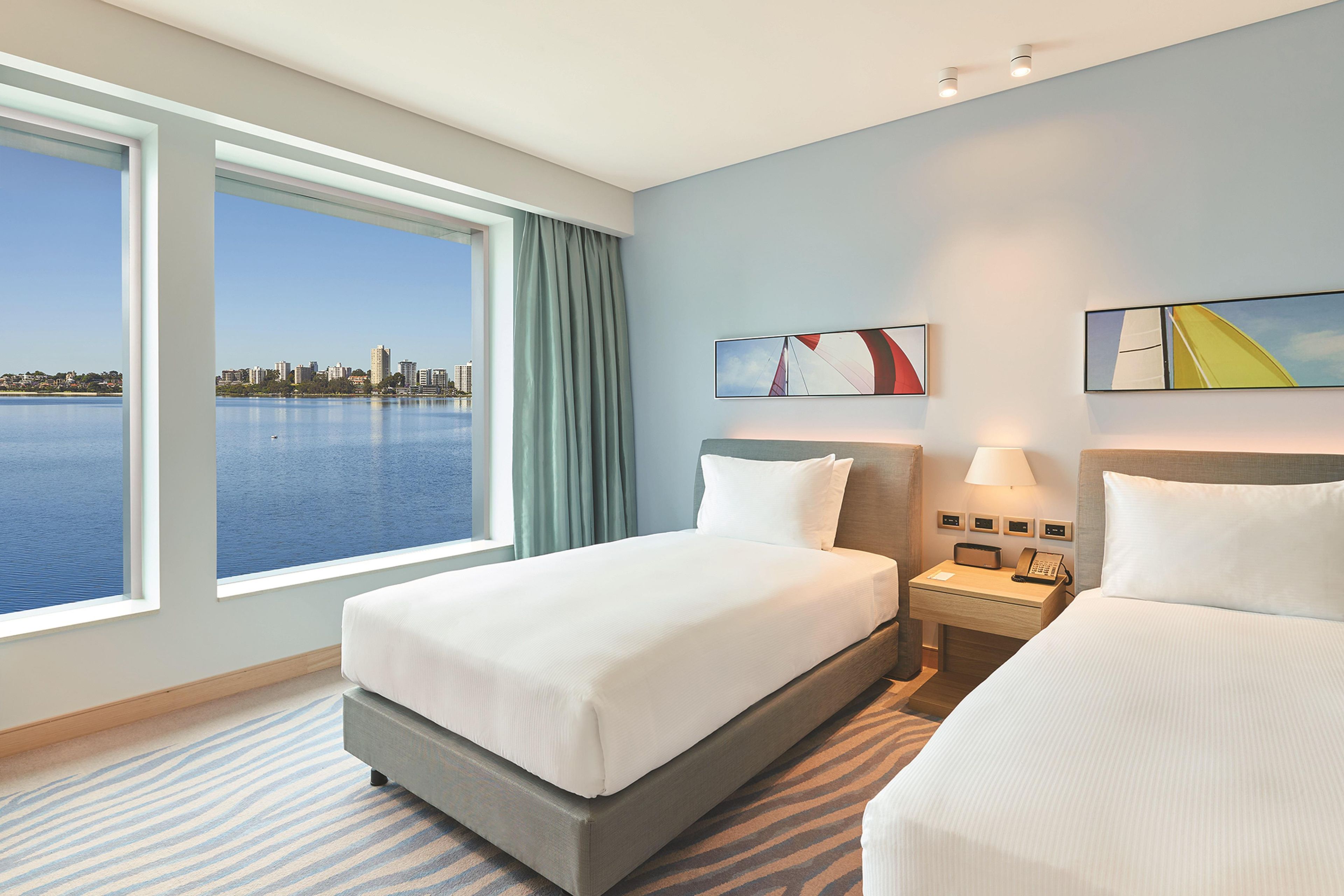 A hotel room with a large window, a bed, a bedside table with a lamp, and a cityscape view.