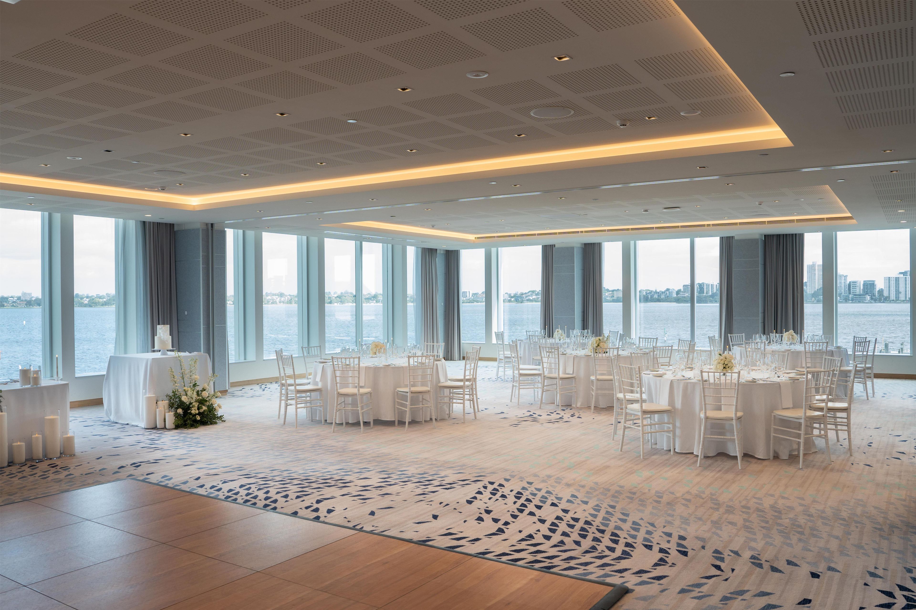 A spacious room with a high ceiling, large windows, and a view of the water. Tables are set for a formal event, and the floor has a patterned carpet.