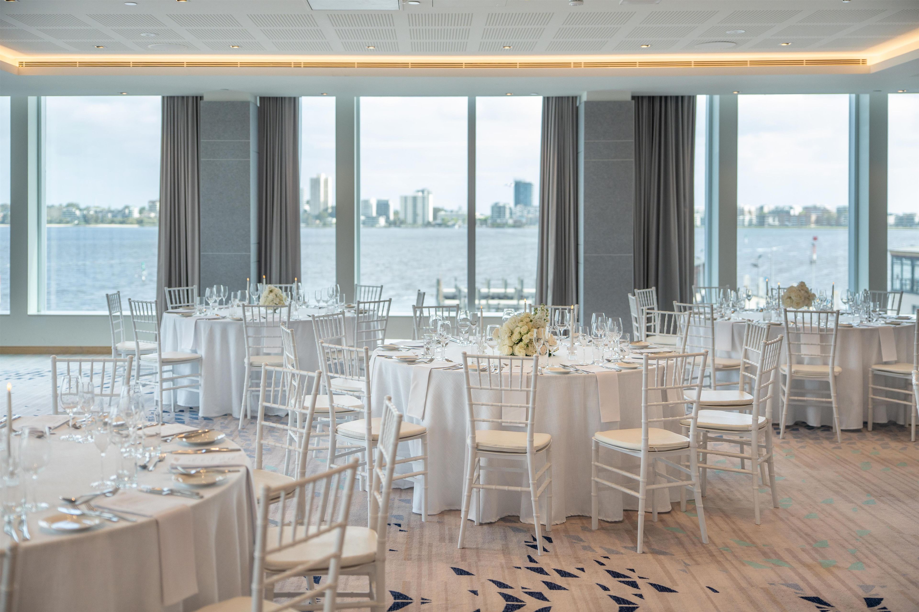 A banquet hall with round tables, white chairs, and a view of a city skyline through large windows.