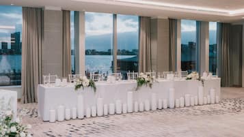 Un salón de banquetes con una larga mesa dispuesta para un evento formal, ventanas amplias con vista a la ciudad y un suelo estampado.