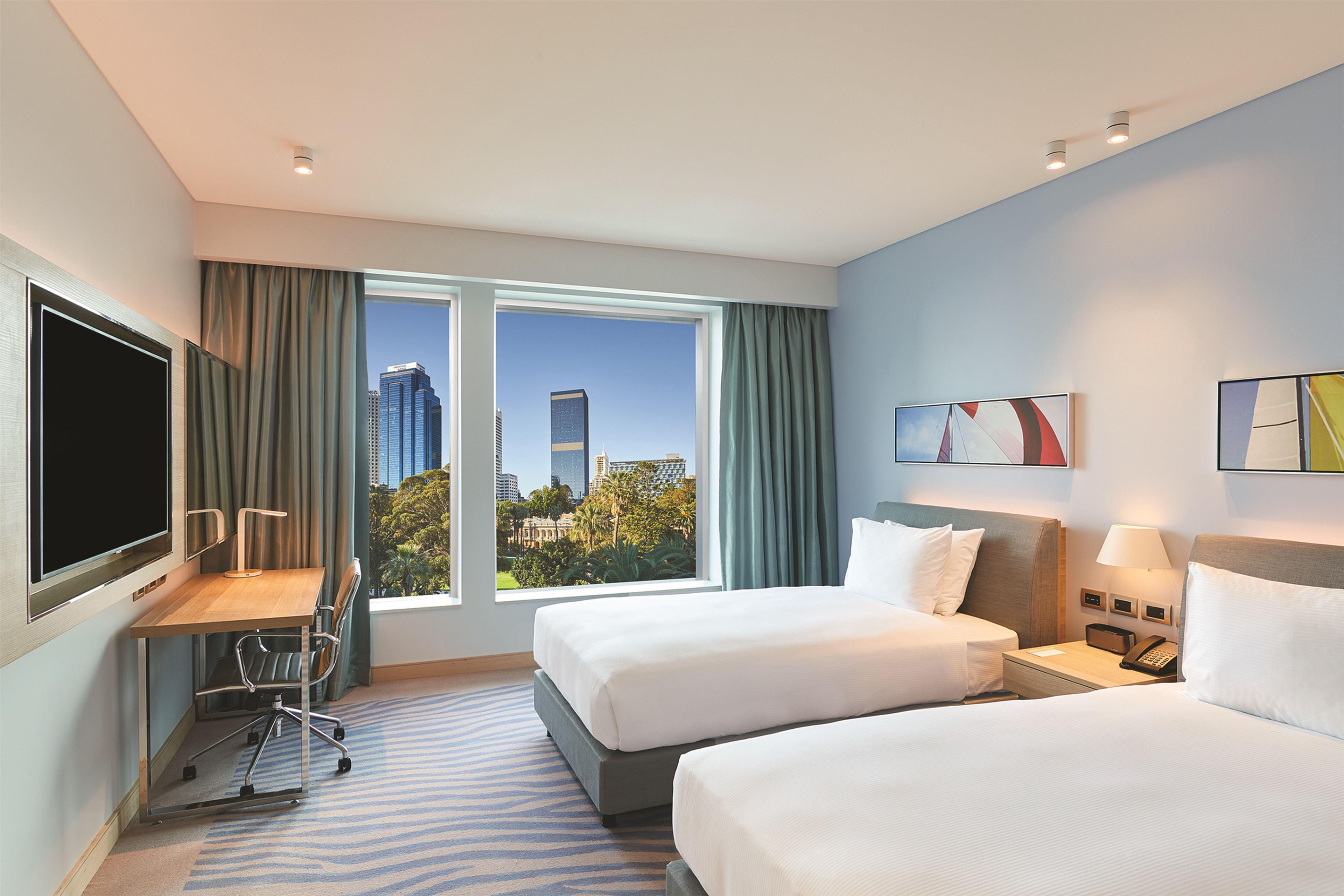 A hotel room with two beds, a desk, a chair, a TV, a window with curtains, and artwork on the wall.