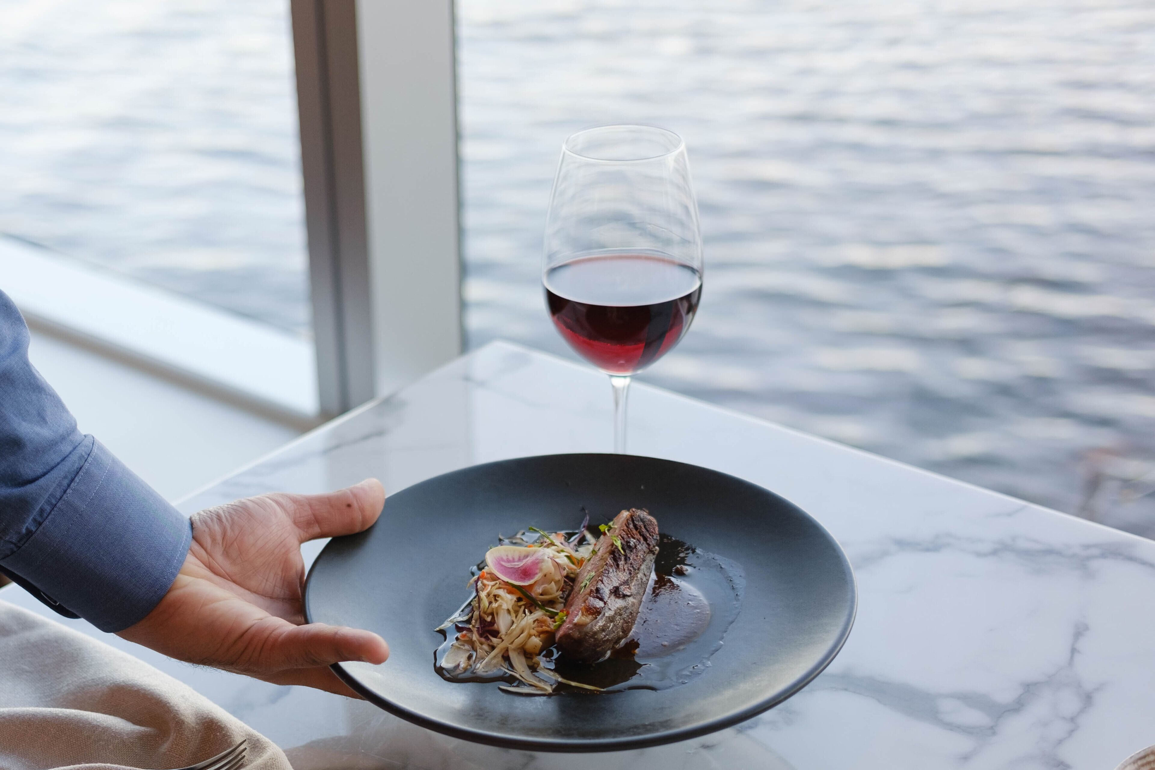 A person holding a plate with a dish and a glass of red wine, with a view of water in the background.
