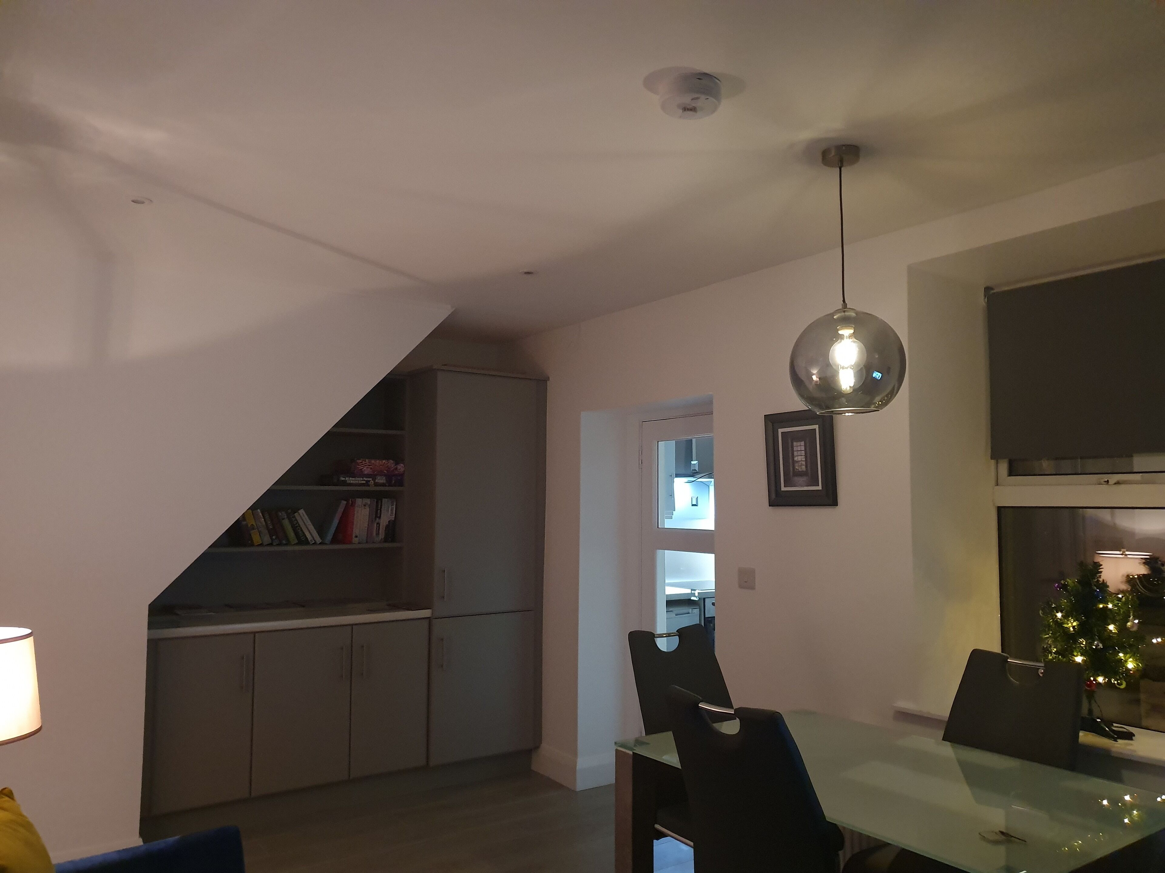 A dining area with a glass table, black chairs, a pendant light, and a bookshelf in the background.
