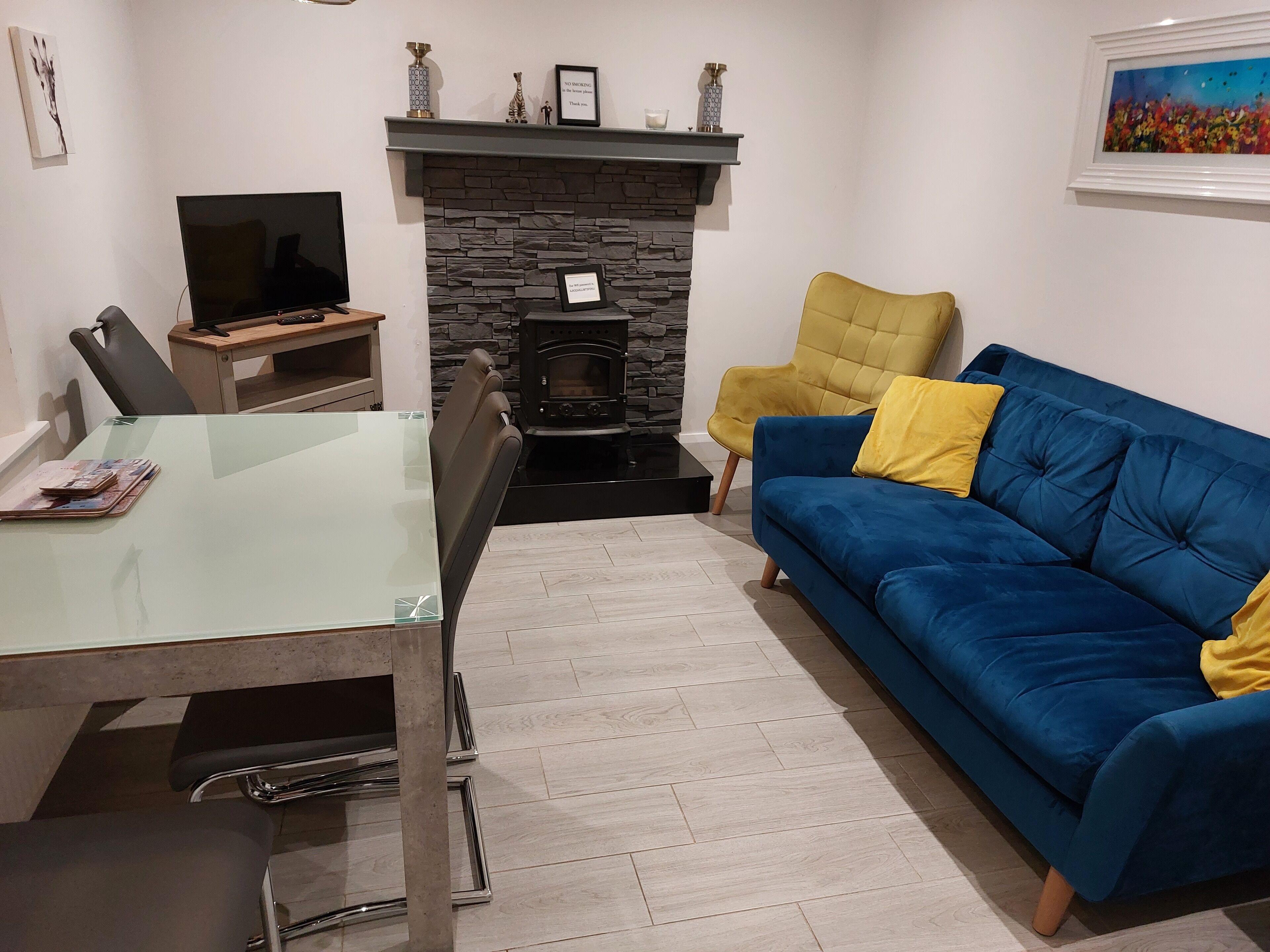 Ein Wohnzimmer mit Kamin aus Stein, einem blauen Sofa mit gelben Kissen, einem Esstisch mit Glasplatte und einem Fernseher.