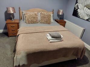 A neatly made bed with a beige blanket, two pillows, and a folded towel on top. There are two bedside tables with lamps, a wooden headboard, and a radiator.