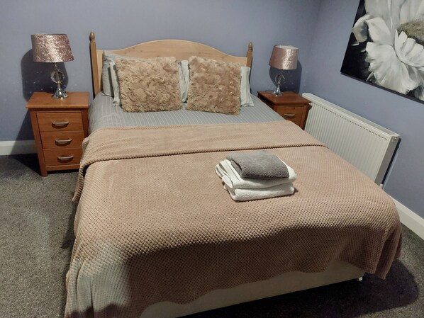 Una cama bien hecha con una manta color beige, dos almohadas y una toalla doblada encima. Se ven dos mesitas de noche con lámparas, un cabecero de madera y un radiador.