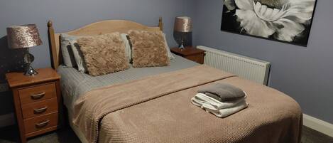 Un dormitorio con cama de madera, dos almohadas mullidas, una mesita de noche con lámpara, un radiador y un cuadro grande con flores en la pared.
