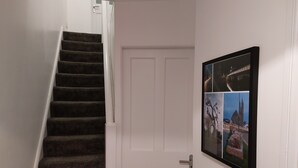 En trappe med mørkt tæppe på trinene, der fører op til en hvid dør. En billedcollage i ramme hænger på væggen til højre.