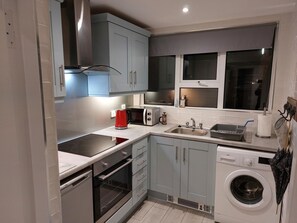 A compact kitchen with grey cabinets, a built-in oven, a washing machine, and a window above the sink.