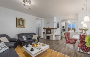 A modern living room with a fireplace, a dining area with red patterned chairs, and a kitchen in the background.