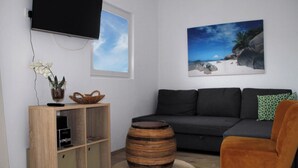 A living room with a dark grey sofa, a wooden cabinet, a round wooden stool, a television mounted on the wall, and a painting of a beach scene. - Holiday home De Wickinger with your own Steiger angler are welcome! (Wervershoof)