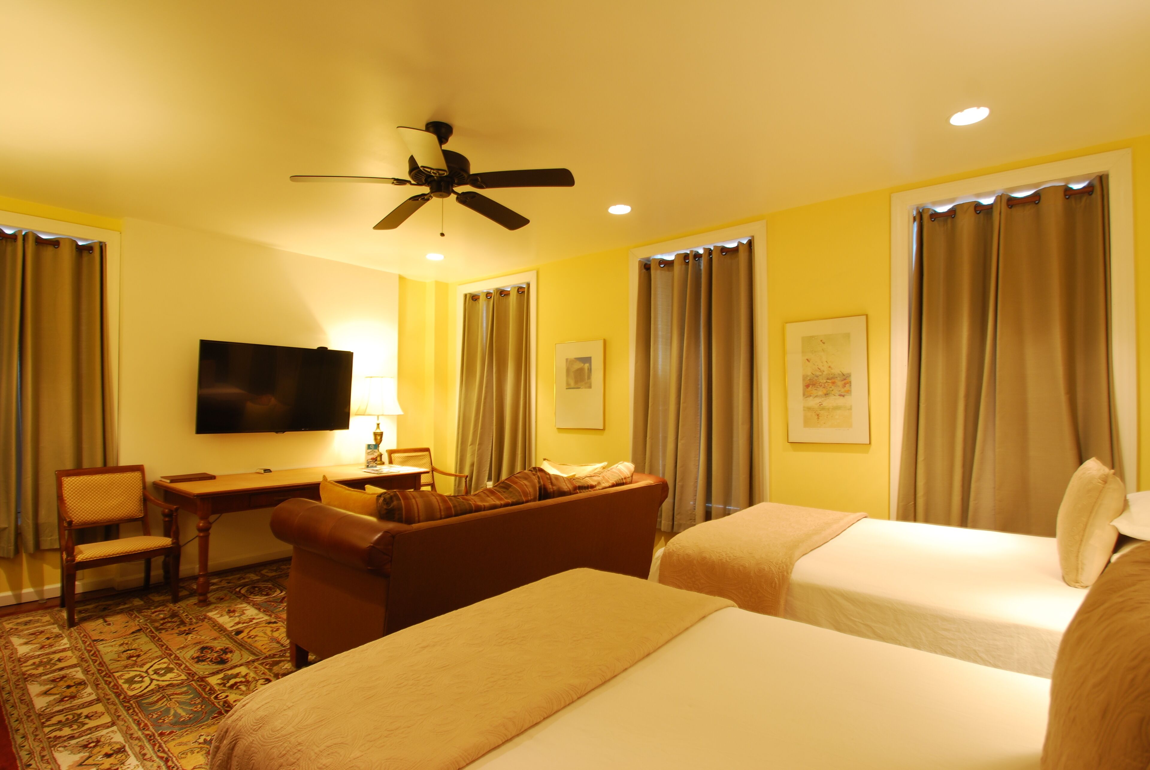 A hotel room with two beds, a desk, a chair, a television, and a ceiling fan.