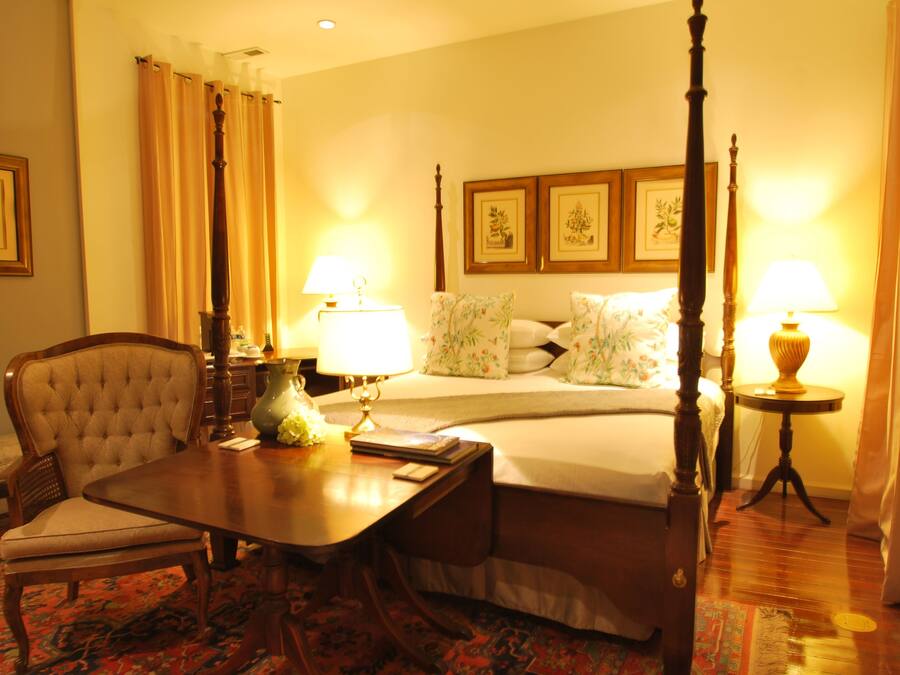 Una habitación de hotel con una cama con dosel, una mesa de comedor con sillas, una mesita de noche con lámparas y una alfombra estampada con motivos florales.