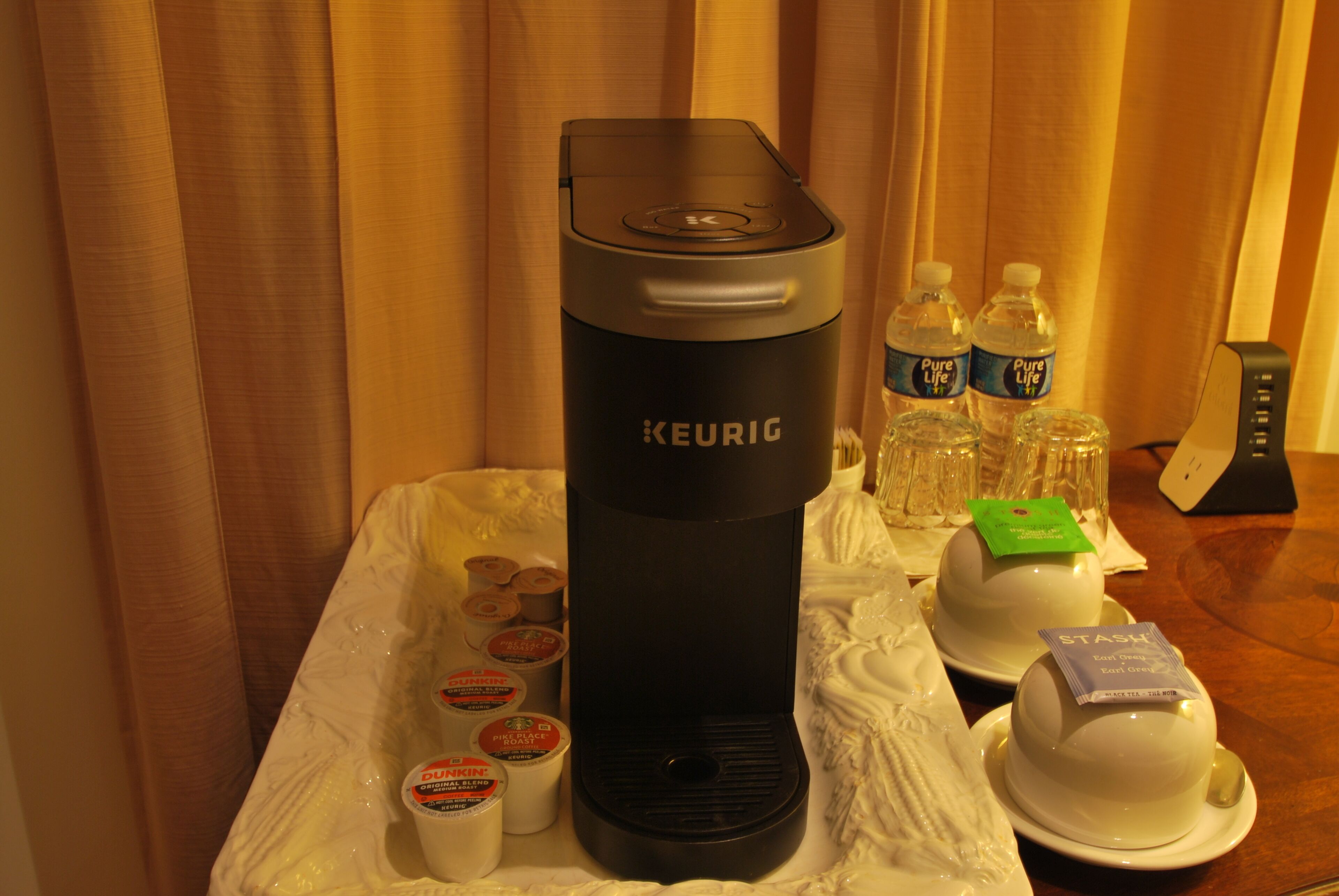A Keurig coffee maker on a table with disposable cups, water bottles, and a tea service.