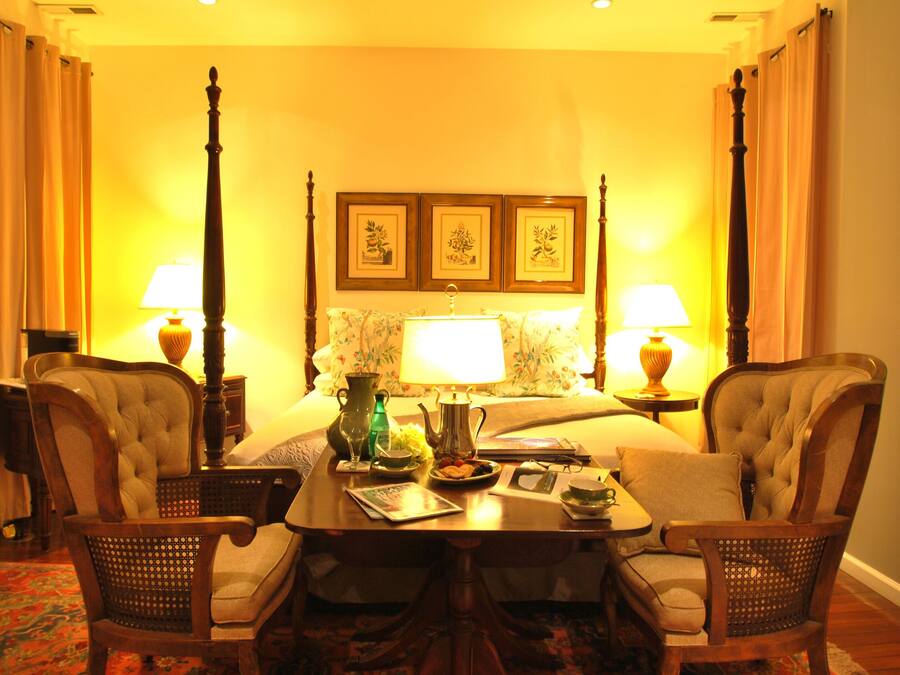 A hotel room with a bed, two armchairs, a table with a lamp, and a bedside table with a lamp. There are framed pictures on the wall above the bed.