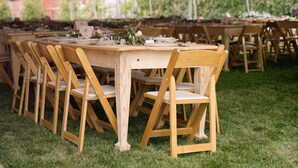 Ein Holztisch, gedeckt für eine Mahlzeit, steht unter einem Zelt mit Lichterketten, umgeben von Stühlen auf einer grünen Wiese.
