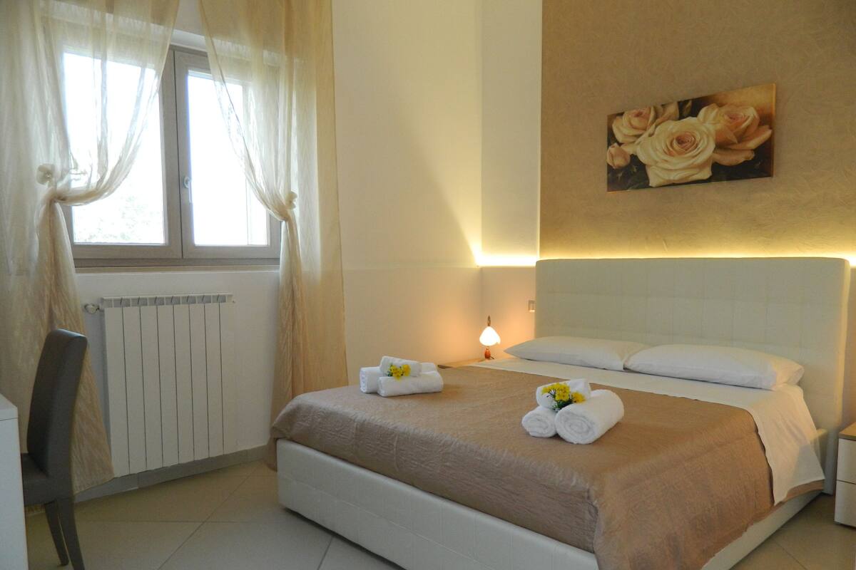 A hotel room with a bed, a chair, a radiator, a window with curtains, and a wall-mounted picture of roses.