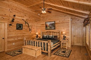 A log cabin bedroom with a bed, two bedside tables, a wooden bench, a ceiling fan, and a painting on the wall.