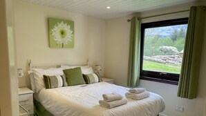 Ein ordentlich bezogenes Bett mit weißen Leinen und grünen Akzenten, ein Nachttisch mit Lampe sowie ein Fenster mit Vorhängen.