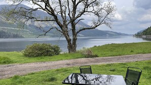 Ein Picknicktisch mit Stühlen und einer Tischdecke, auf einer Terrasse mit Blick auf einen See und Berge.