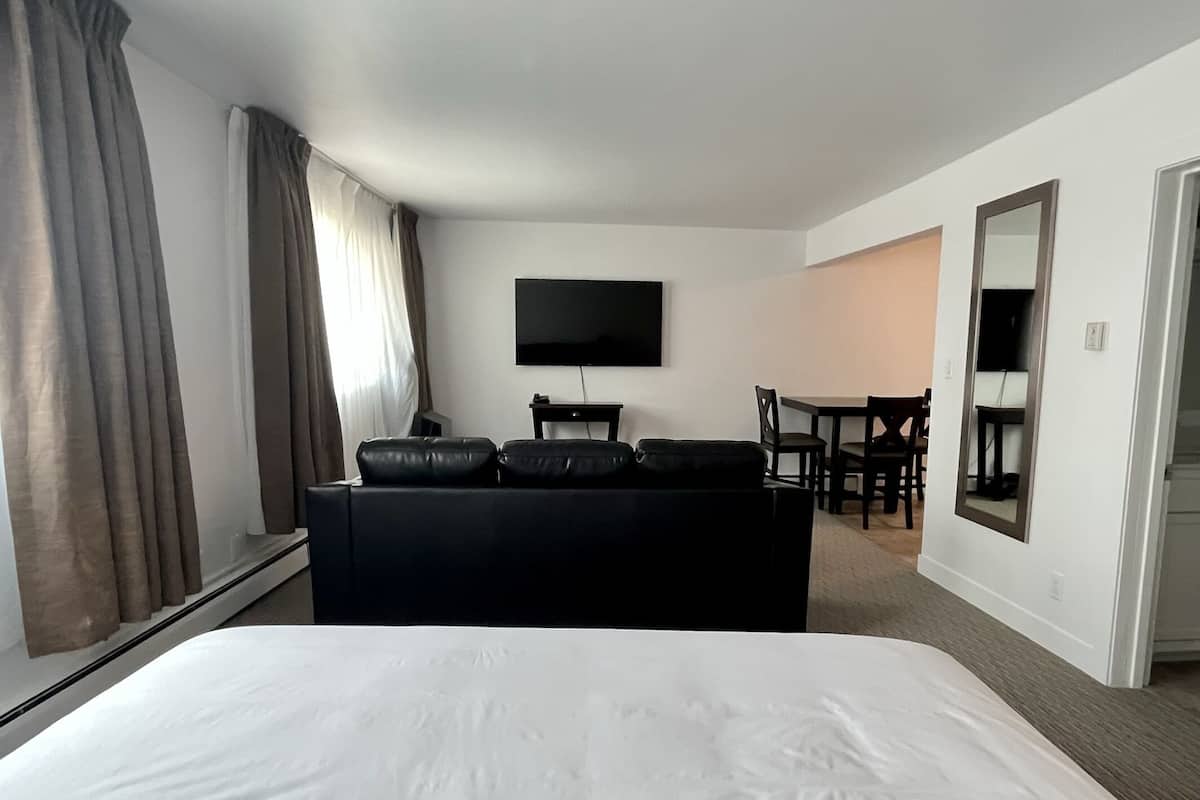 A hotel room with a large bed, a black sofa with cushions, a flat-screen TV mounted on the wall, a dining table with chairs, and a mirror on the wall.