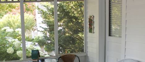 A room with a glass table, two wicker chairs, and a large window with a view of trees and a parked car.