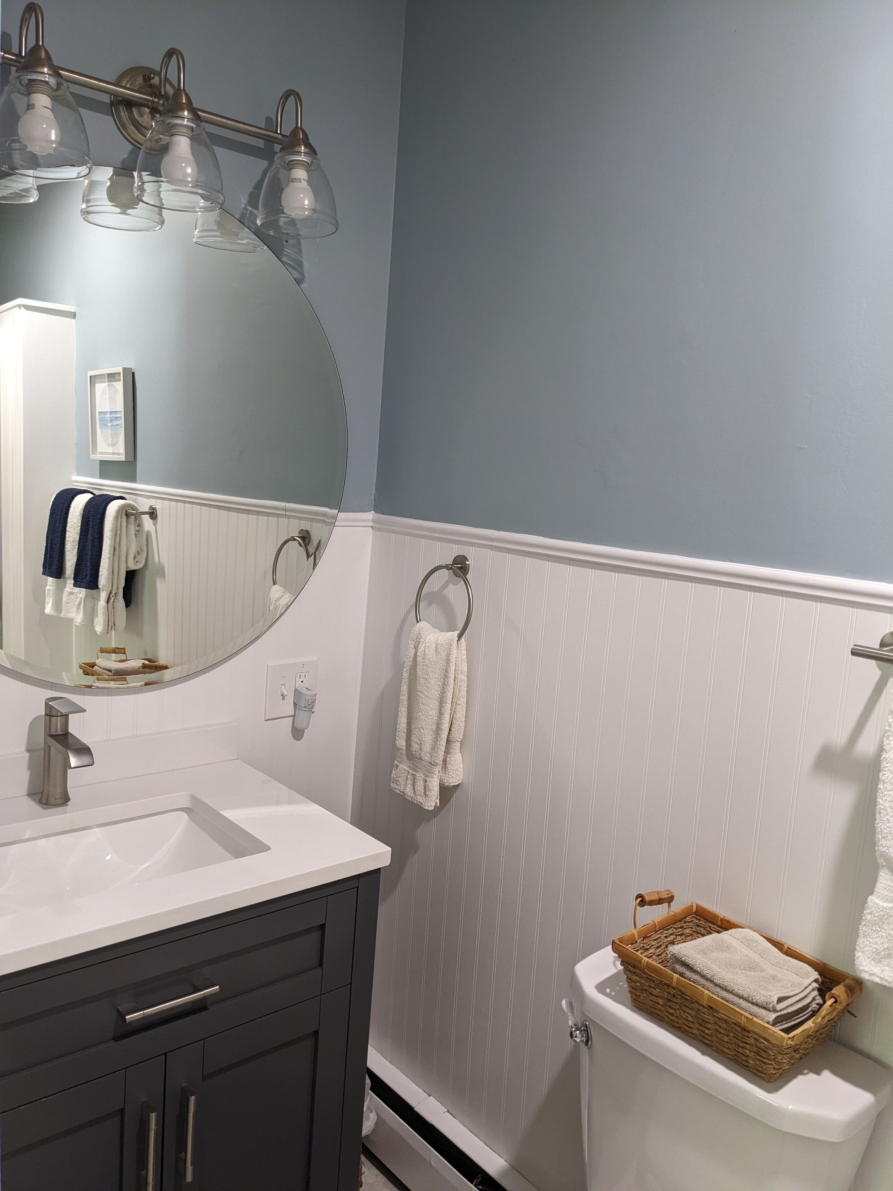 A bathroom with a white sink, dark cabinets, a round mirror, and a toilet with a basket of towels.