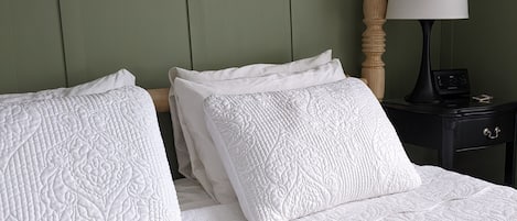 Een netjes opgemaakt bed met een gewatteerd sprei en kussens, een houten hoofdbord, een nachtkastje met een lamp en een klok, en een groene muur.