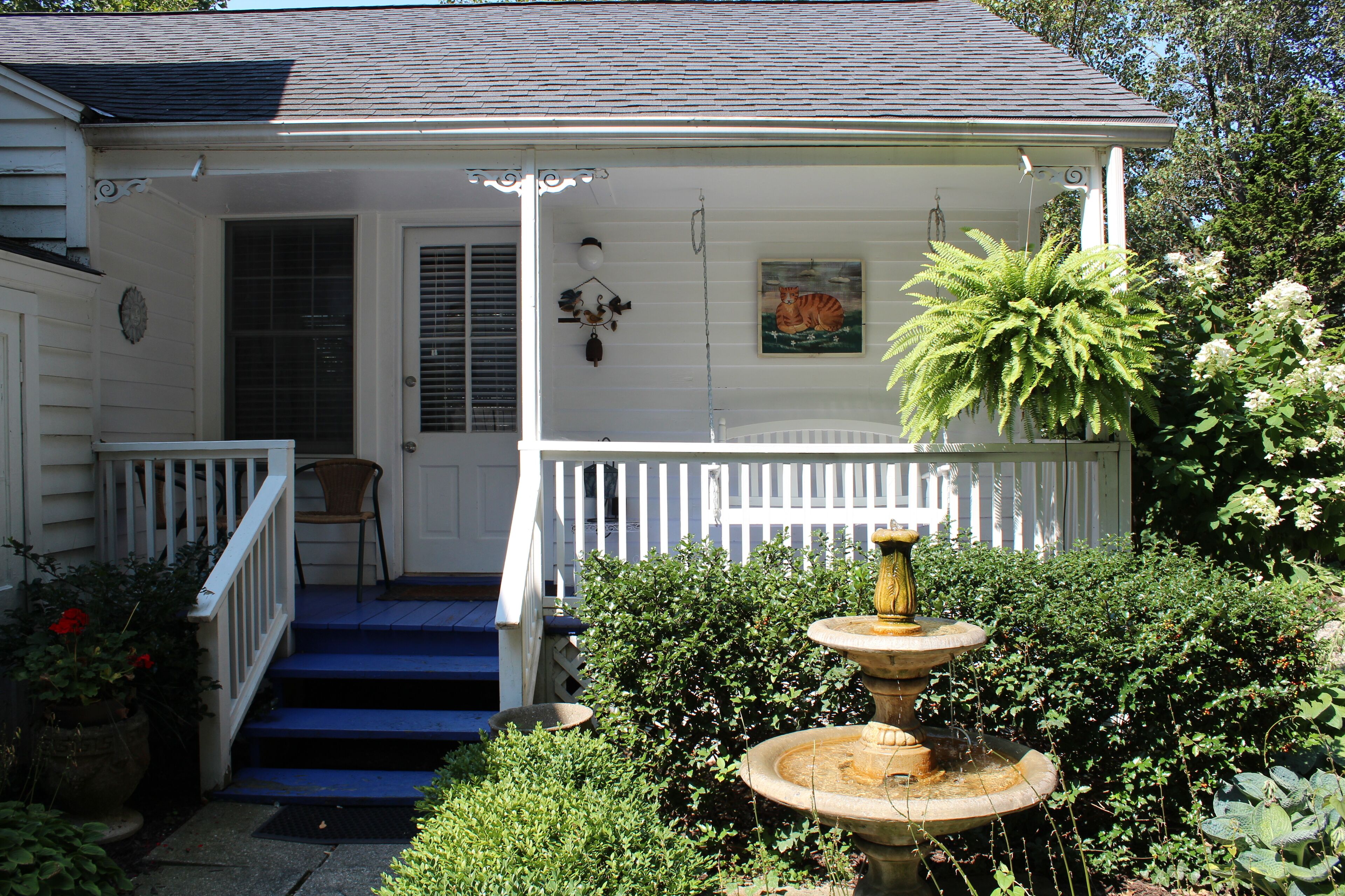 A white house with a porch, blue steps, a white railing, a door with shutters, a painting of a tiger, and a fountain.