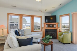 A living room with a fireplace, a television, and large windows. - Life Is Good: Ocean & Sound Views, Private Pool & Hot Tub, Quick Walk to Beach (Nags Head)